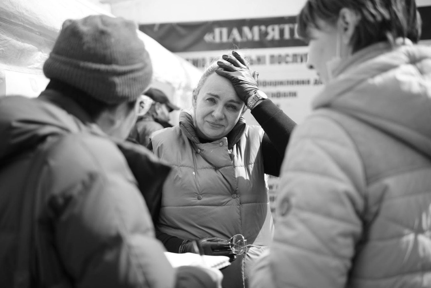 Чорно-біле фото: жінка в жилеті та рукавичках біля намету з інформаційним стендом у Бучі